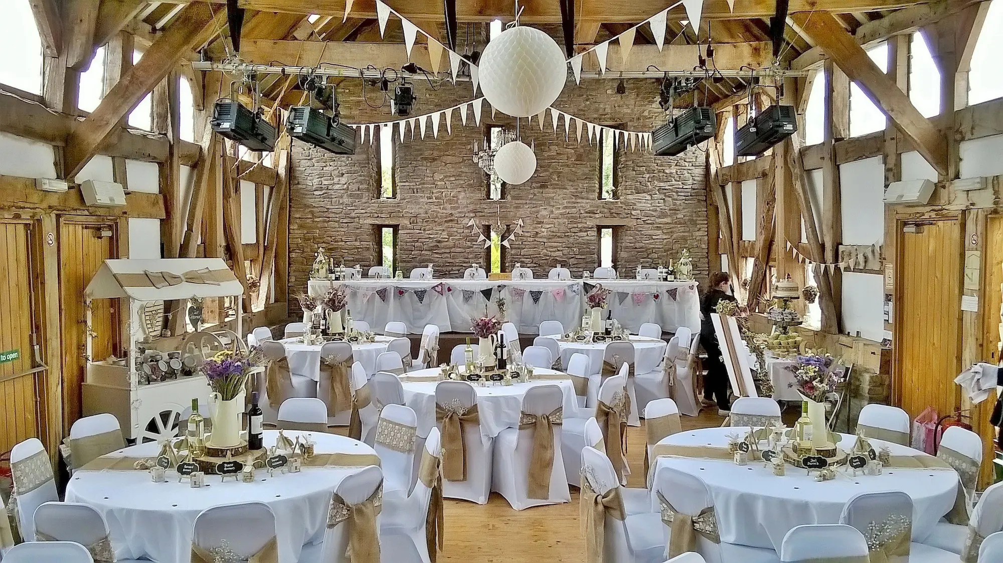 The airy interior of a converted timber-framed barn, decked with celebratory bunting as if for a wedding. The floor is filled with multiple circular tables, decked in white. At the end of the room, there is a long rectangular table on a raised platform.
