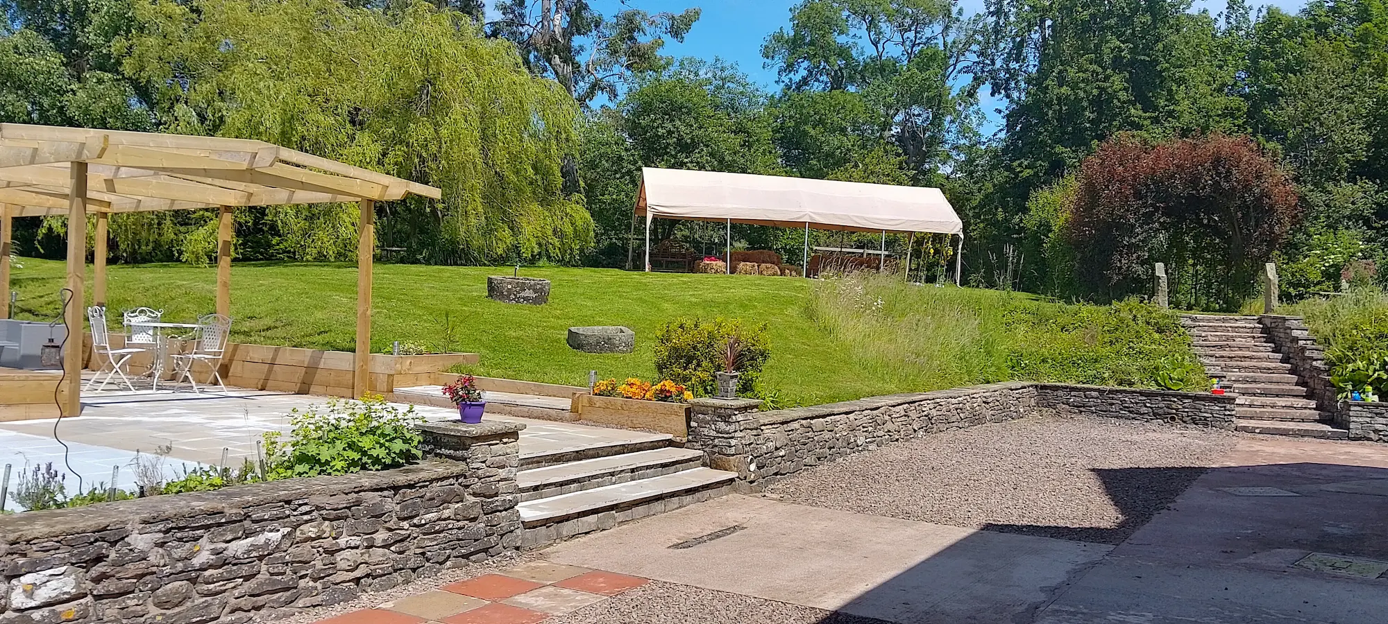 A sunlit garden. Steps lead up from a gravelled patio area to a sitting area next to green lawn. On the other side of the lawn, a gazebo stands by some trees.